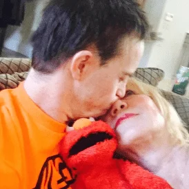 A person in an orange shirt kisses a woman on the cheek while holding a red Elmo toy, symbolizing the warmth and love central to the Christopher Smith Foundation's mission of caregiver support and recognition within the disability community.| A Non Profit For Caregivers | Christopher Smith Foundation