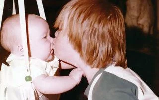 A young child gently kisses a baby, who is seated in a swing, highlighting a tender moment of caregiving support. The Christopher Smith Foundation celebrates such acts of love and dedication in the disability community.| A Non Profit For Caregivers | Christopher Smith Foundation