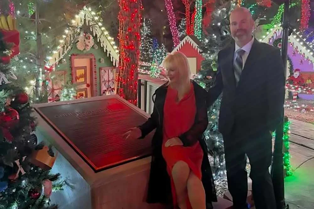 A man in a suit and a woman in a red dress stand beside a plaque surrounded by festive lights and trees, celebrating caregiver recognition at a Christopher Smith Foundation event.| A Non Profit For Caregivers | Christopher Smith Foundation